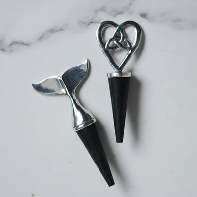 Two rubber wine bottle stoppers with pewter heads, one is a whale tail and the other is a celtic knot in the shape of a heart, on a light gray background.